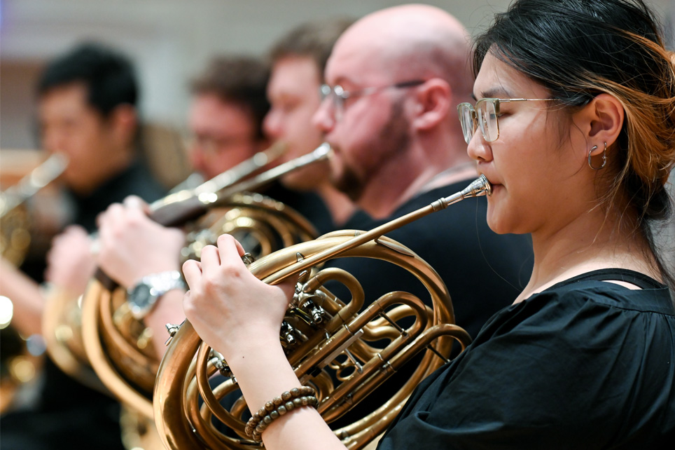 A line of students, wearing black clothes, playing the French horn, in a brass ensemble.